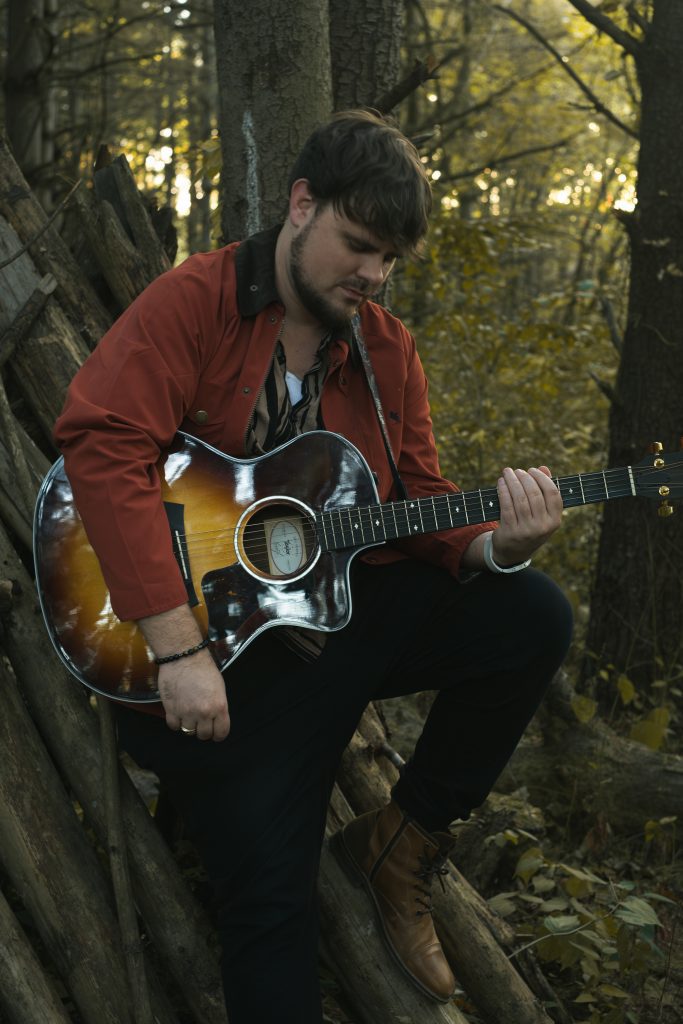 Bio photo - Heath Church playing guitar and sitting by a tree in the woods
